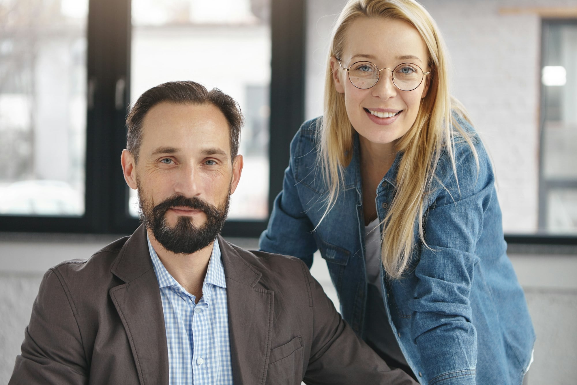Confident bearded male enterpreneur and secretary sit close to each other in office, look confidentl